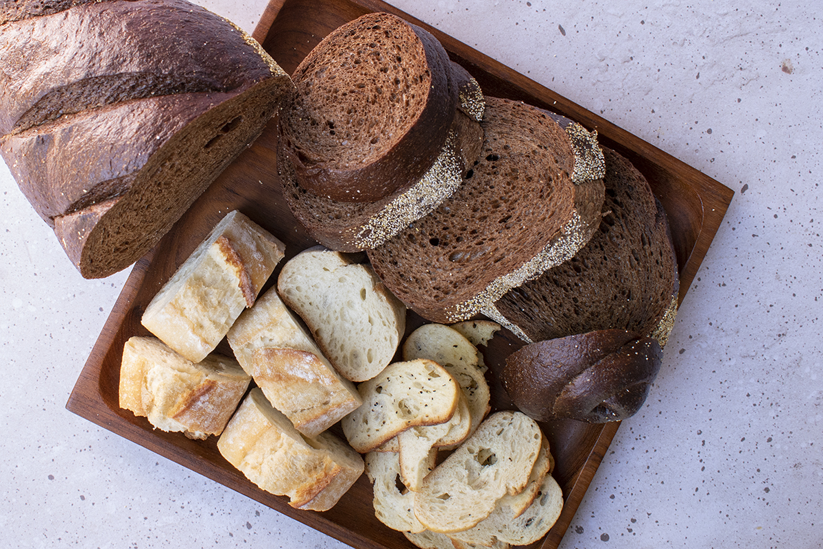 Overhead view of different types of bread on a wood platter, pumpernickel, baguette and crostini.