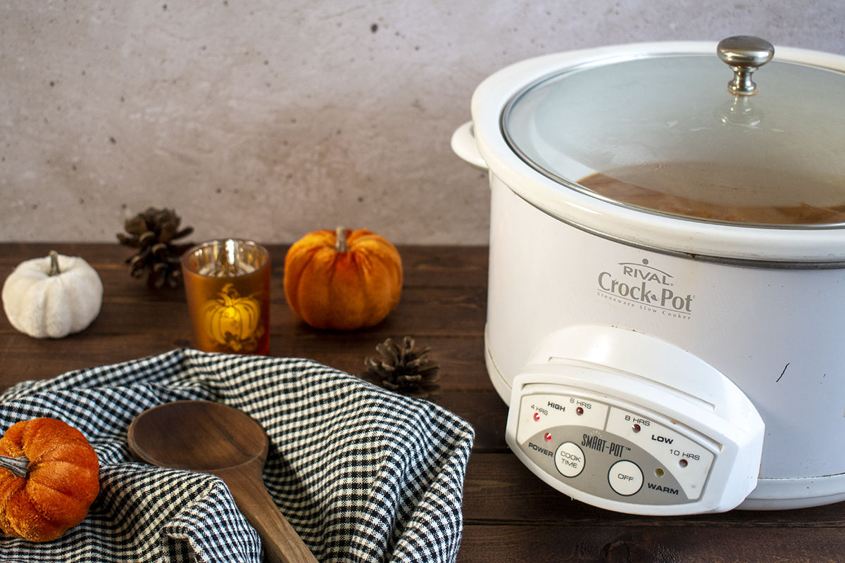 Slow cooker set up on a table with fall decor and a wooden spoon set on a black and white gingham kitchen towel.