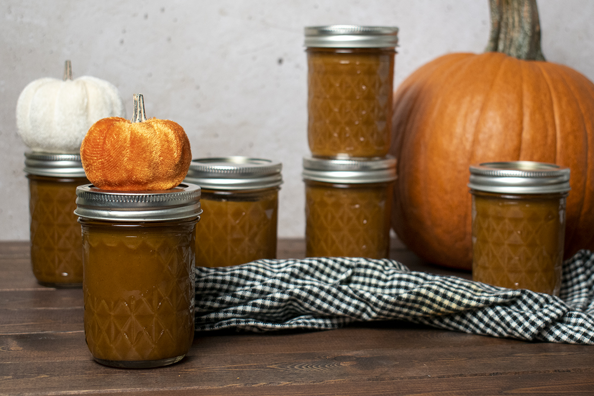 jars of pumpkin butter with a pie pumpkin in the background, a black and white gingham kitchen towel and decorative velvet pumpkins in the foreground.
