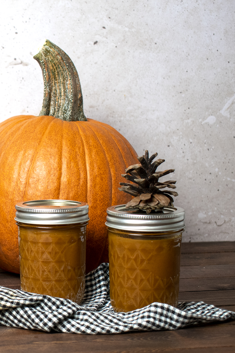 Two jars of pumpkin butter sitting on a black and white gingham kitchen towel, pie pumpkin in the background 