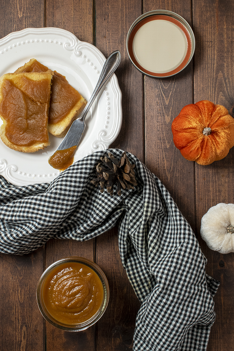 Overhead view of a jar of pumpkin butter, toast spread with pumpkin butter with small decorative pumpkins and a black and white kitchen towel 