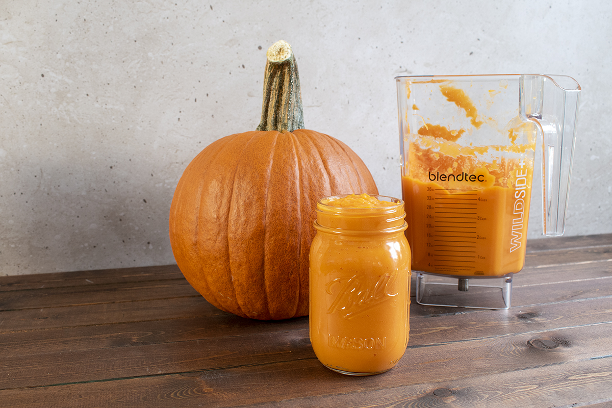A jar of pumpkin puree in front of a pie pumpkin and blender jar with pumpkin puree. 