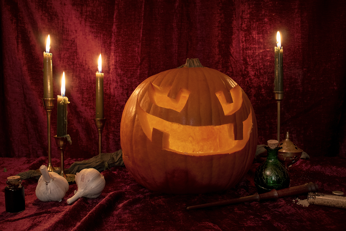 Large jack-o-lantern lit up, on a velvet covered table surrounded by candles, potions, garlic and a wand.