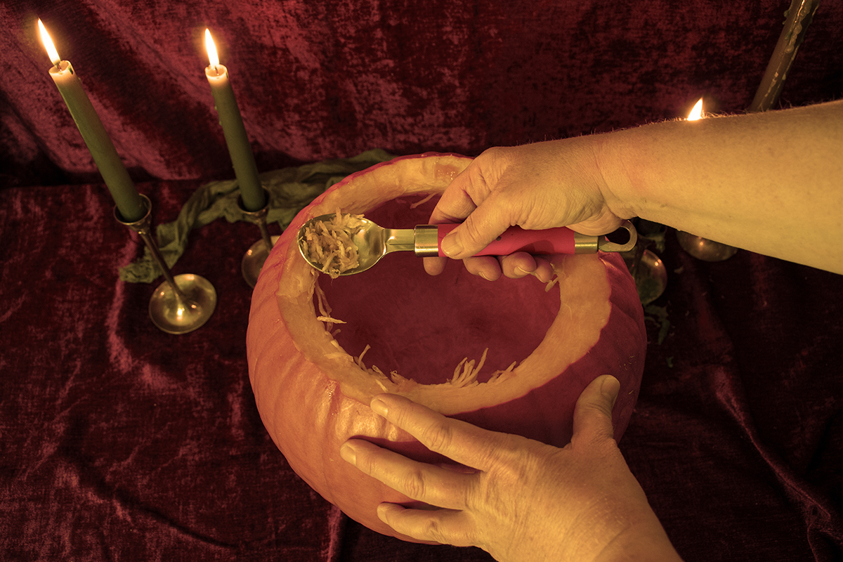 Woman's hands holding the side of a pumpkin and using an ice cream scoop to scoop out the flesh.