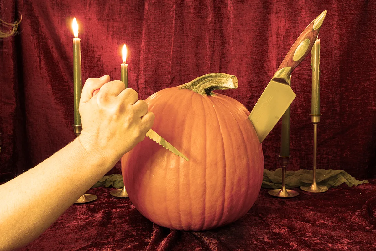 A large kitchen knife sticking out of the side of a pumpkin and a female hand holding a small serrated pumpkin knife in front of the pumpkin.