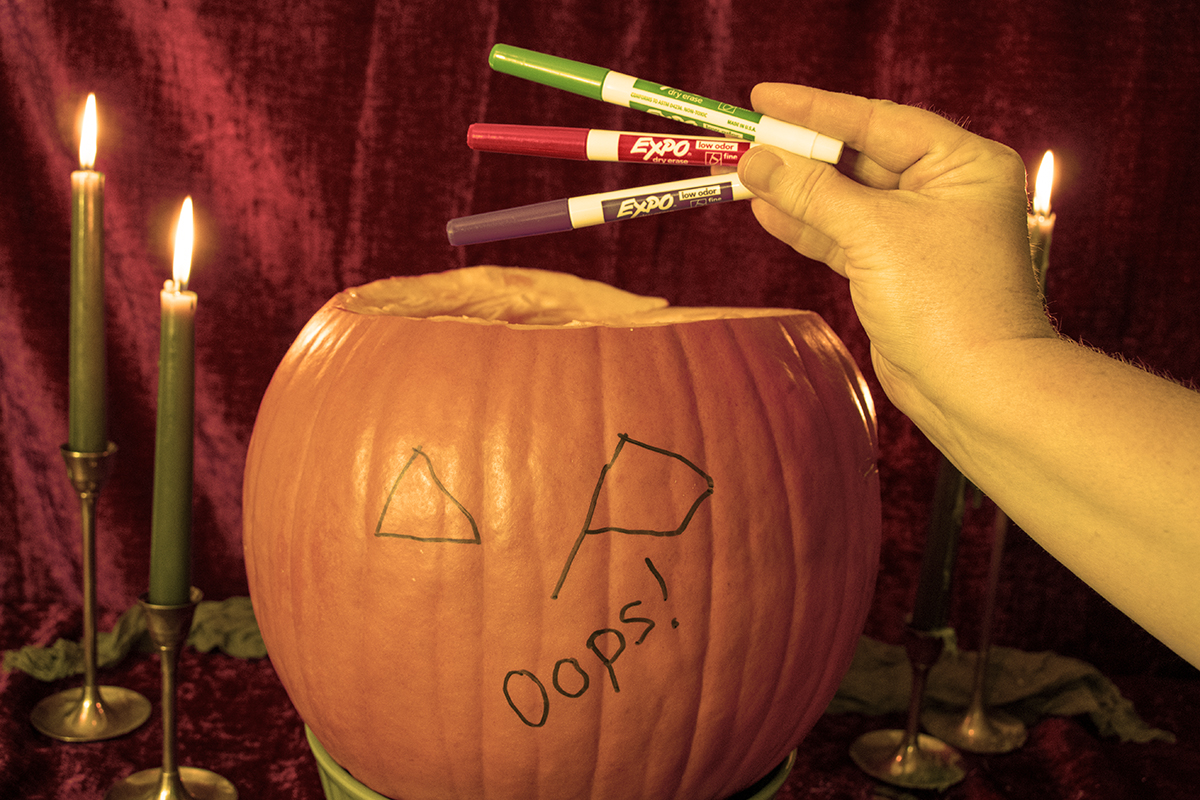 A pumpkin with writing on it, a messed up face and the words 'oops' and a woman's hand holding up three dry-erase markers.