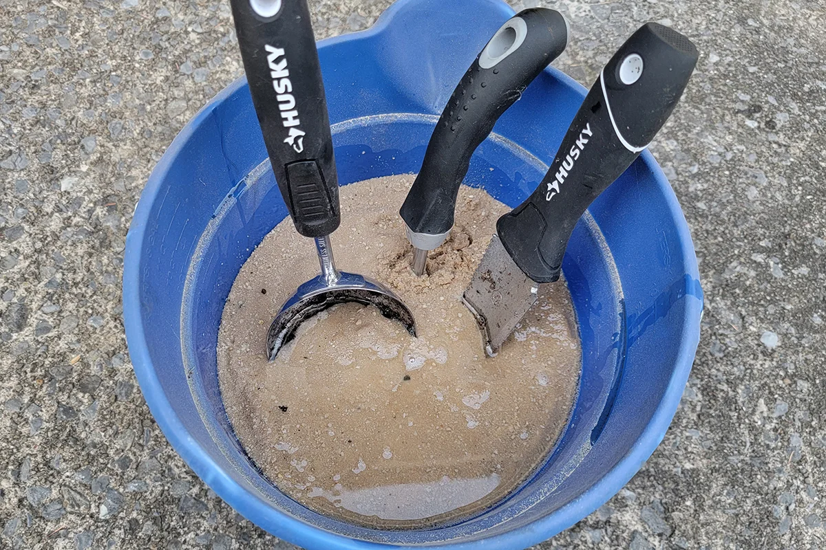 Bucket filled with sand and mineral oil with garden tools stuck in the sand.