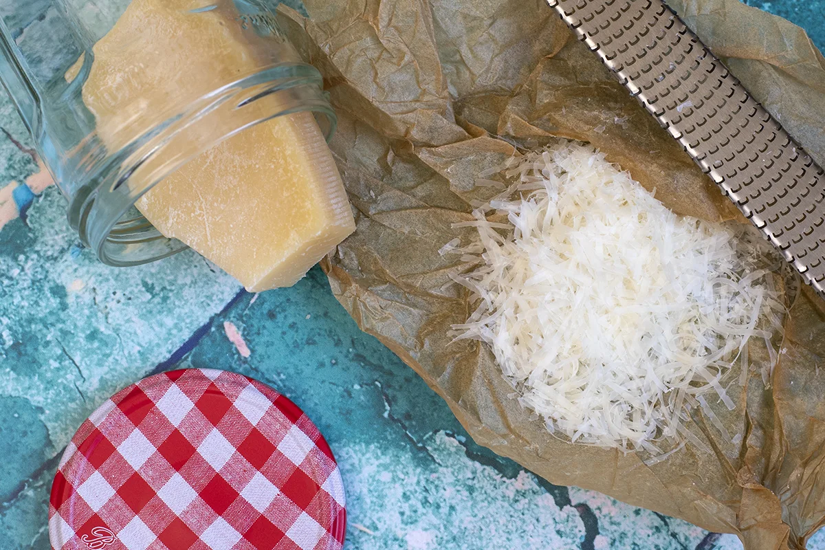 Grated parmesan cheese on parchment paper and a chunk of parmesan cheese in a clear glass jar.