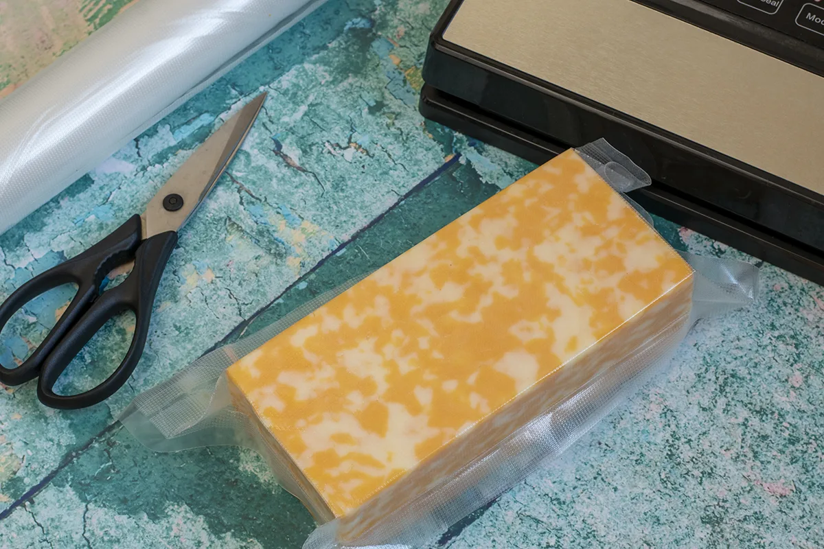 A large block of Colby Jack cheese vacuum-sealed, sitting next to the vacuum sealer, scissors and plastic roll.
