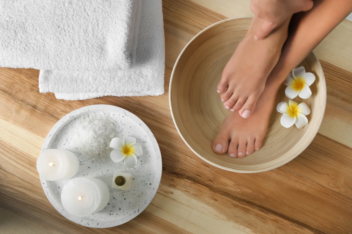 Female feet soaking in a basin of water, candles and flowers nearby with a fluffy white towel. 