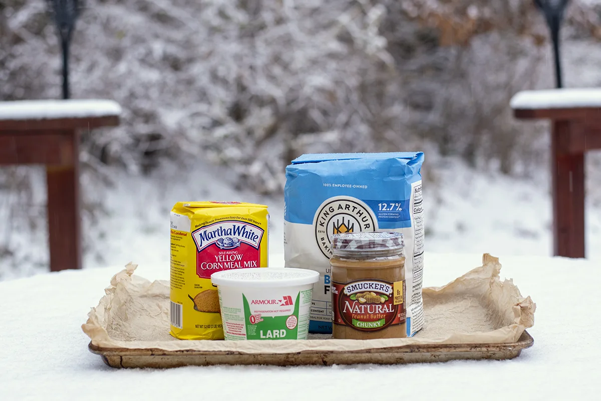Suet cake ingredients sitting on a parchment-lined baking sheet in the snow. A tub of lard, a jar of peanut butter a sack of flour and a small sack of cornmeal.