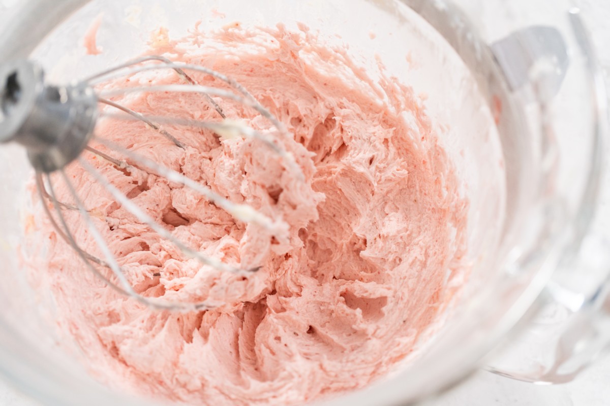 Overhead view of beater bowl with strawberry buttercream frosting, whisk to the side.