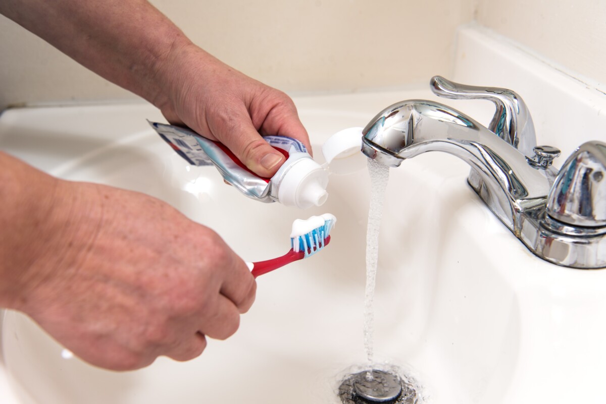 Hands shown putting toothpaste on a toothbrush while the water runs from the tap.
