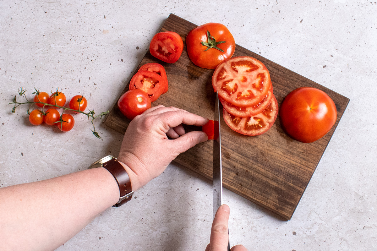 Woman's hands slicing a cherry tomato on a cutting board with a chefs knife.