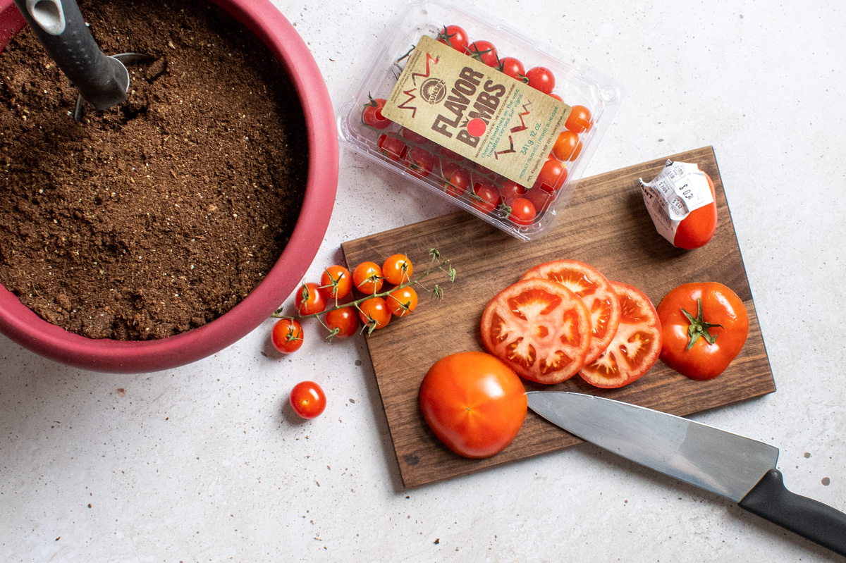 Overhead shot of pot of soil next to a cutting board with several types of sliced tomatoes. 