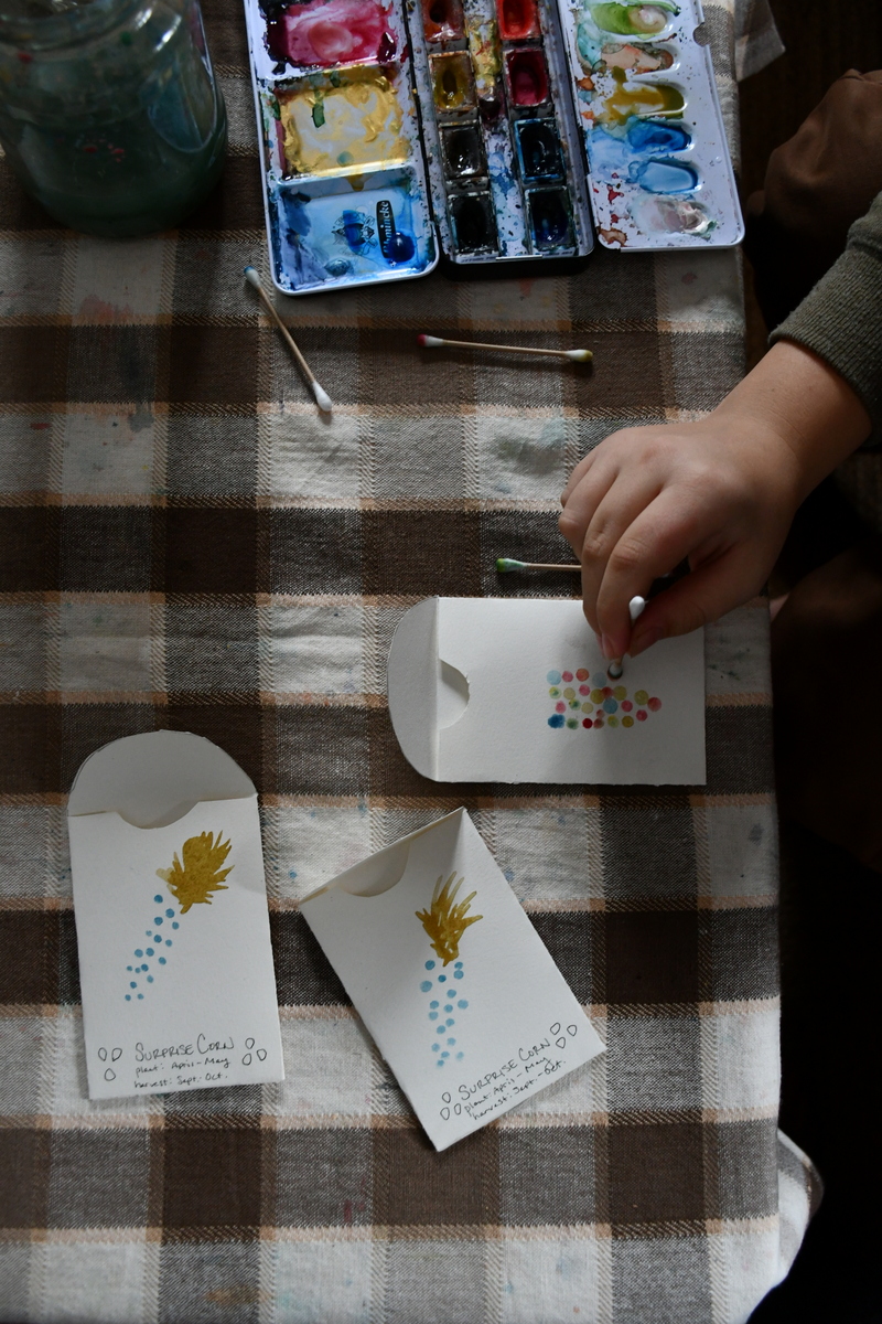 Child's hand using a cotton swab to create image of corn on seed packet