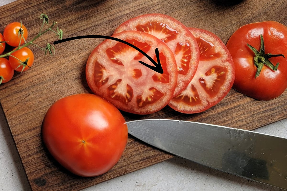 Sliced tomato on a wood cutting board next to a knife. Black arrow graphic pointing to a tomato slice.