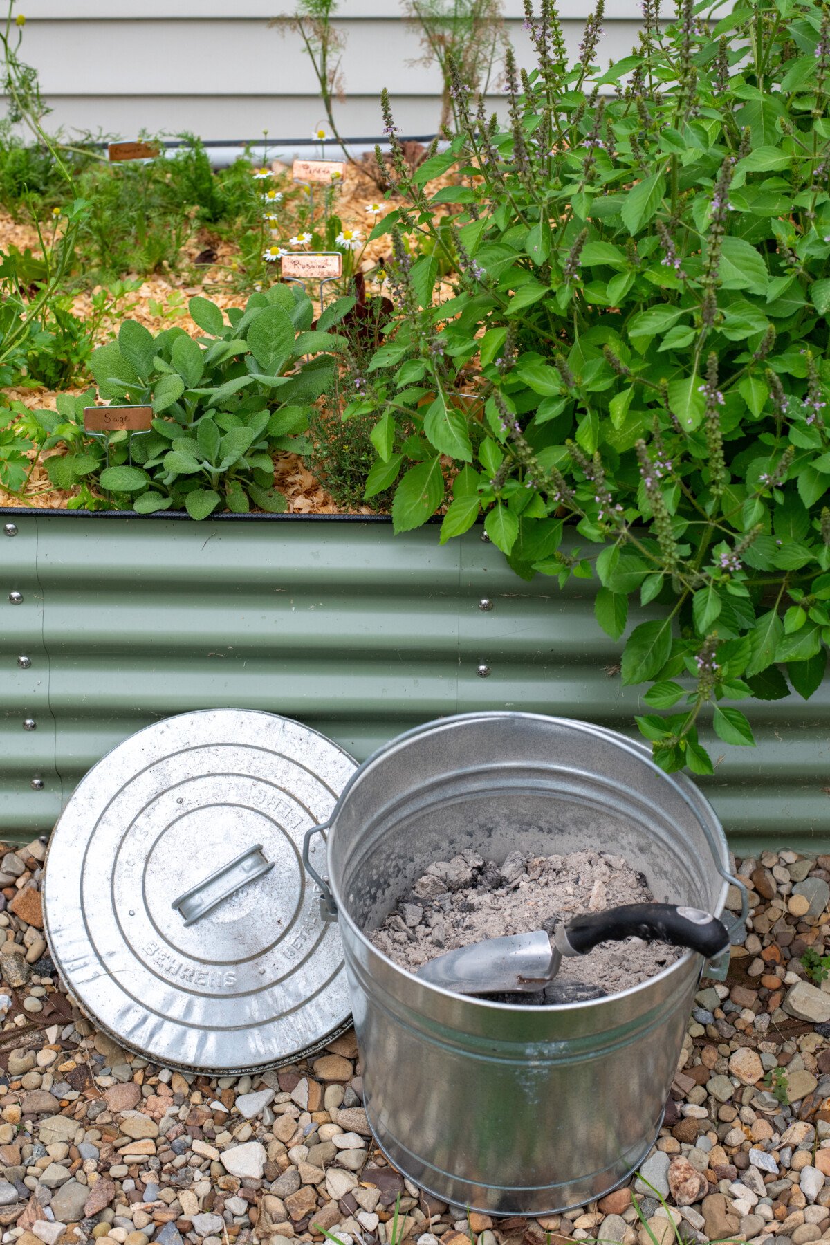 Metal bin filled with wood ash, lid next to it, next to raised bed garden