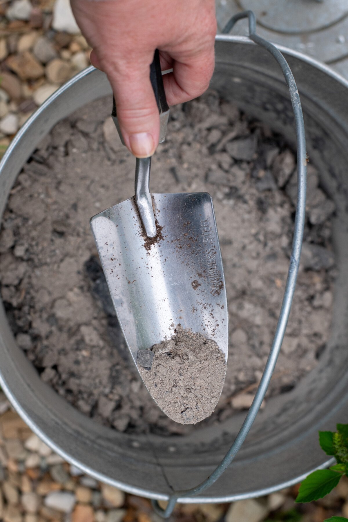 Hand holding spade with wood ash over bucket