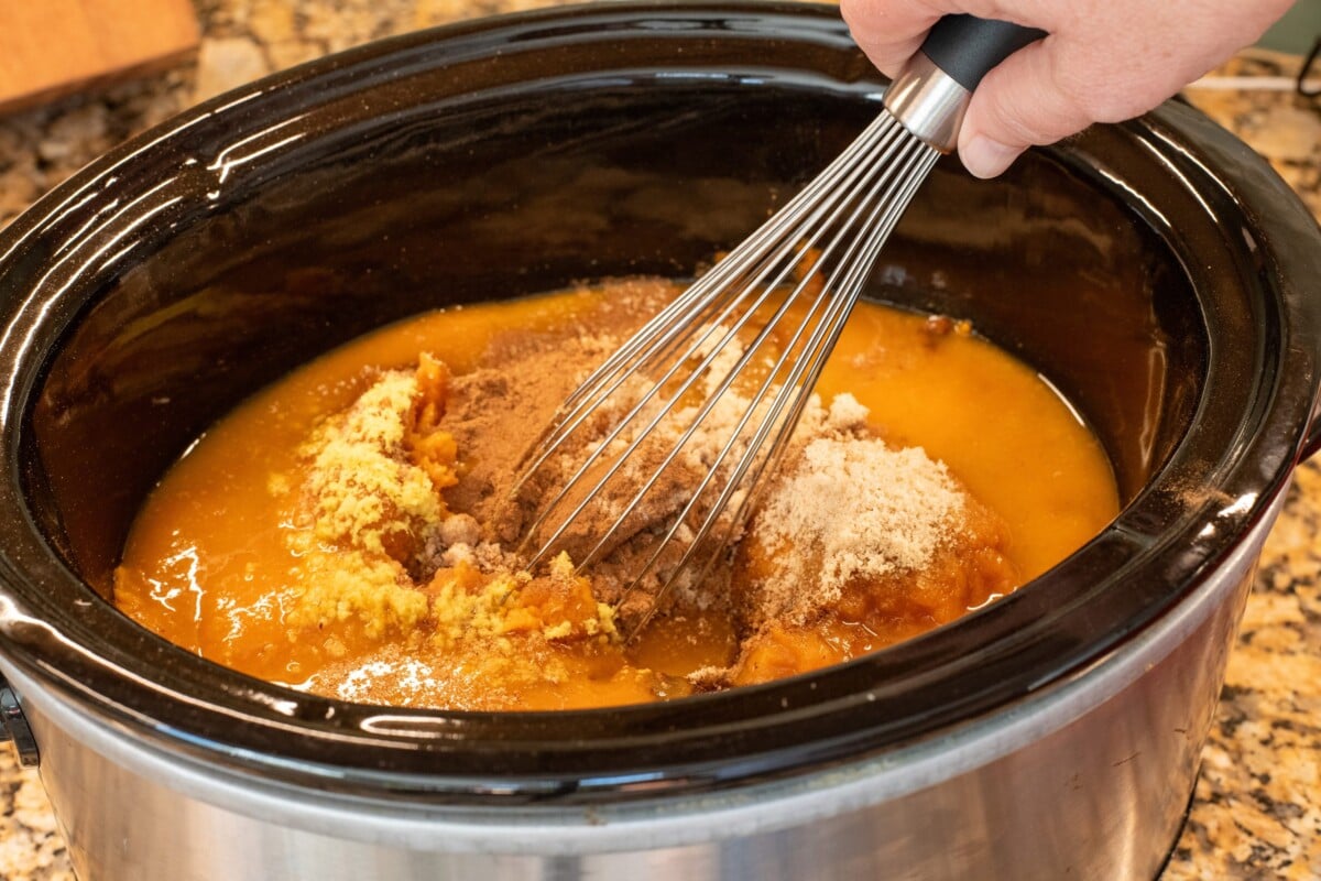 Woman's hand stirring pumpkin butter ingredients in a slow cooker with a whisk.